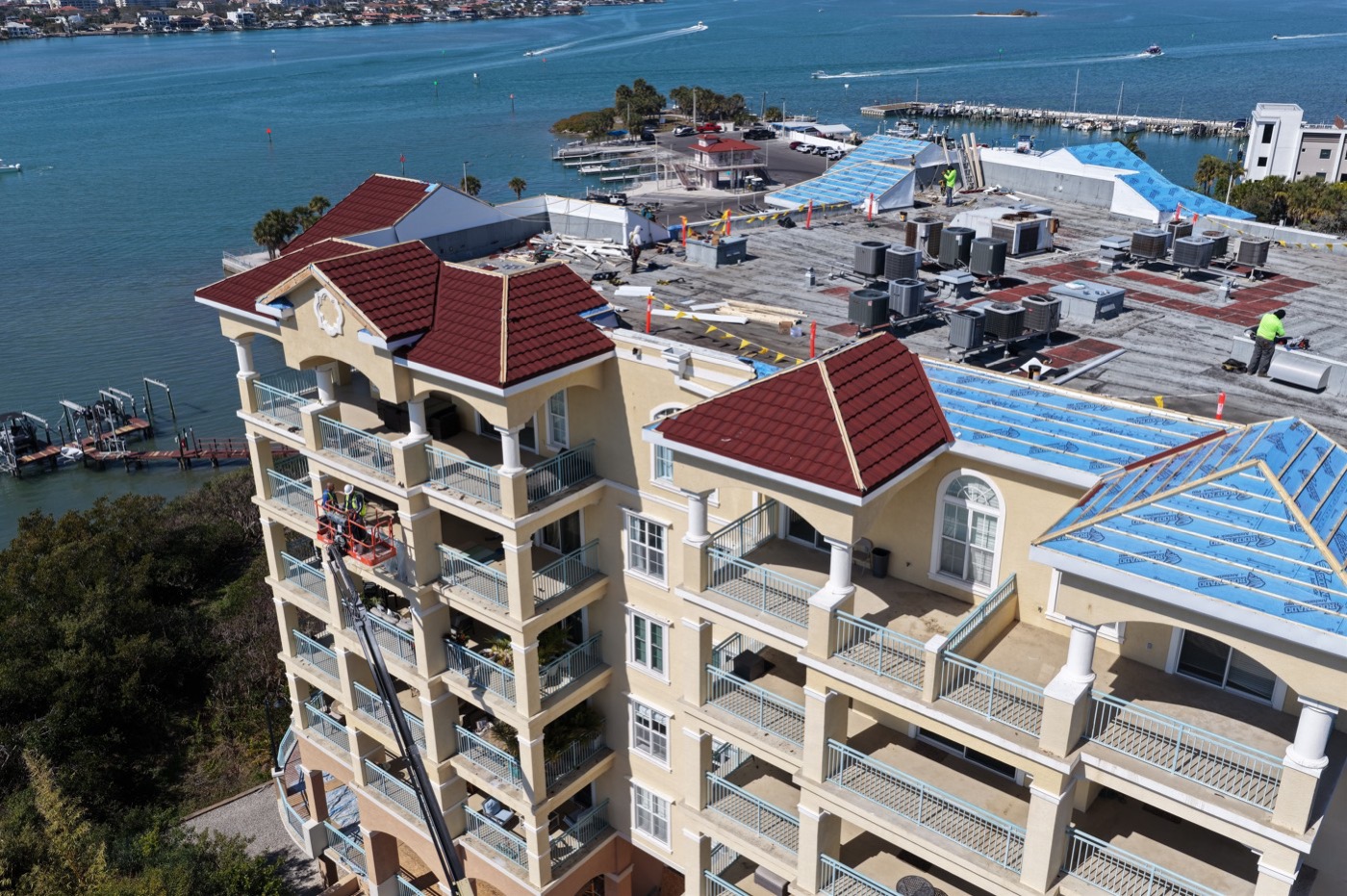 Aerial view of roof work in progress on a waterfront condo building, with crews and a lift working on the new tile roof